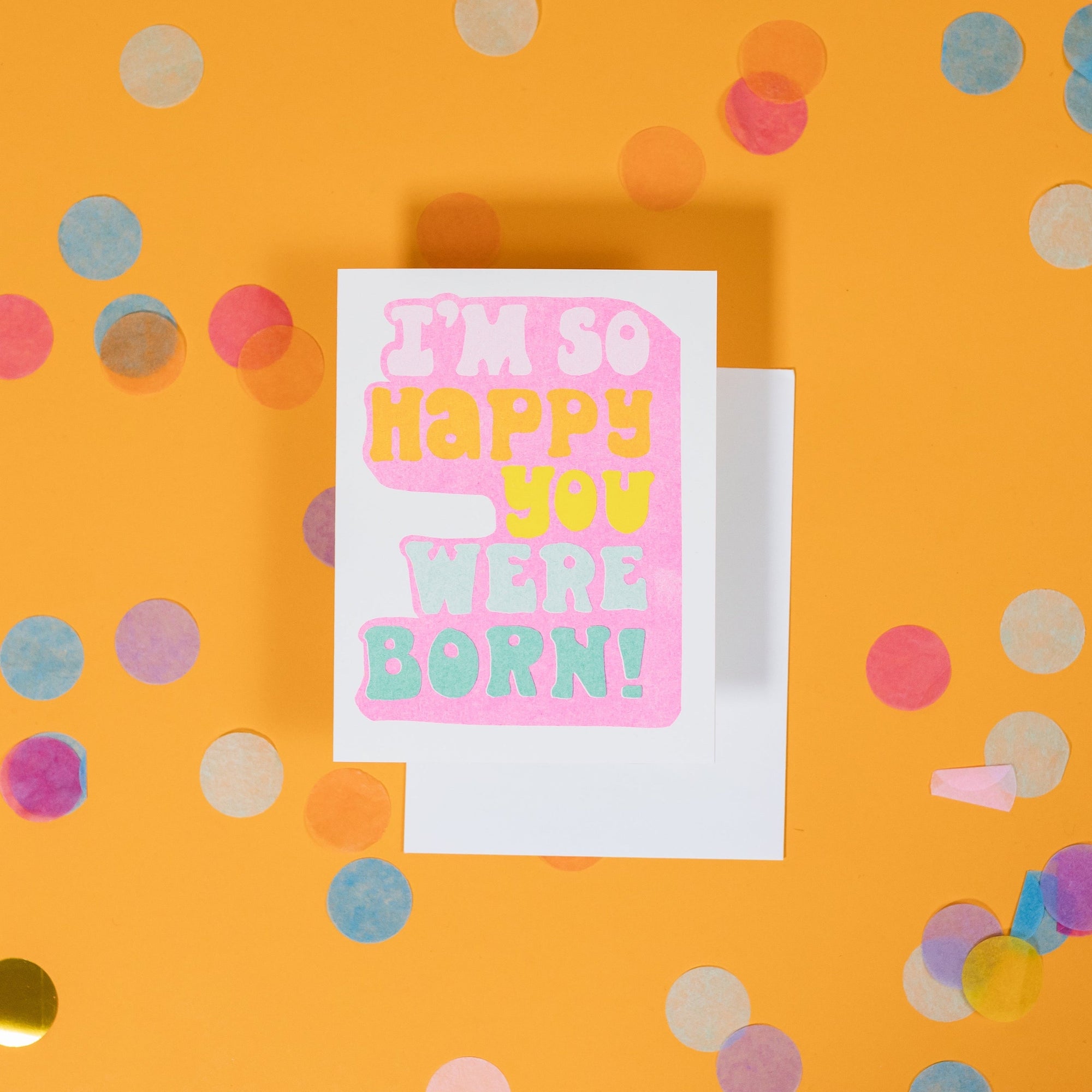 On a sunny mustard background is a greeting card and envelope with big, colorful confetti scattered around. The white greeting card says "I'M SO HAPPY YOU WERE BORN!" in handwritten funky lettering in white, orange, yellow, cool aqua, turquoise with pink underneath. The white envelope sits under the card. 