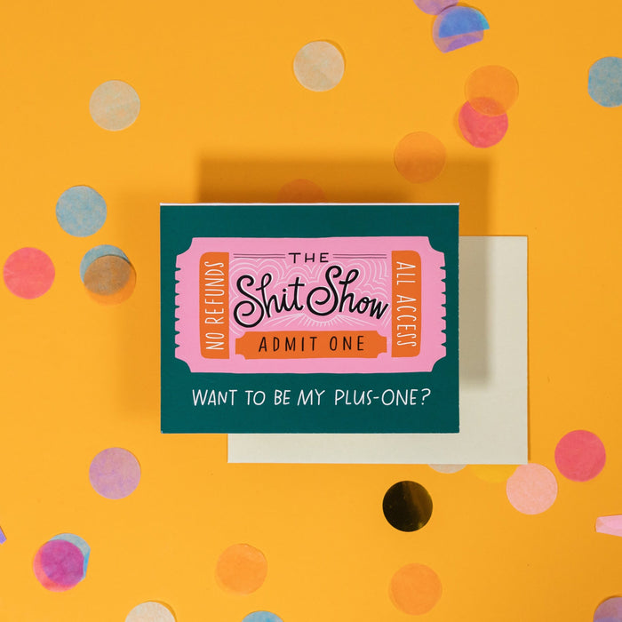On a sunny mustard background is a greeting card and envelope with big, colorful confetti scattered around. The dark emerald green greeting card has an illustration of a bubblegum pink and orange ticket on it and says "The Shit Show Admit One", "No Refunds All Access", "Want To Be My Plus-One?" in handwritten white and black lettering. The light green envelope sits under the card. 