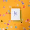 On a sunny mustard background is a greeting card and envelope with big, colorful confetti scattered around. The white greeting card has an illustration of a pope sitting on the toilet and it says "HOLY SHIT!" in handwritten purple lettering. The chartreuse envelope sits under the card. 