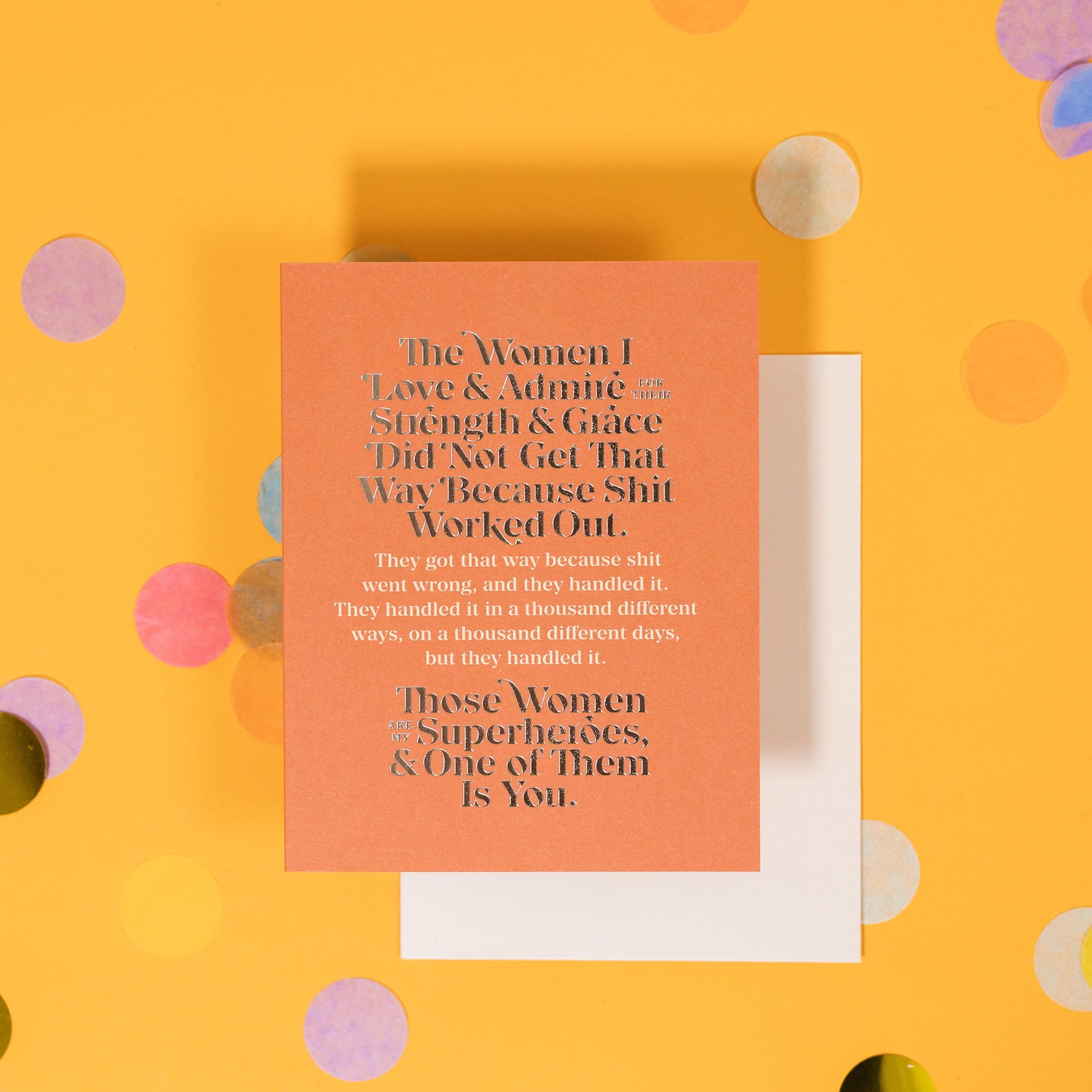 On a sunny mustard background is a greeting card and envelope with big, colorful confetti scattered around. The orange greeting card says "The Women I Love & Admire For Their Strength & Grace Did Not Get That Way Because Shit Worked Out. Those Women Are My Superheroes, & One of Them Is You." It is written in gold foil and white serif font. The white envelope sits under the card. 