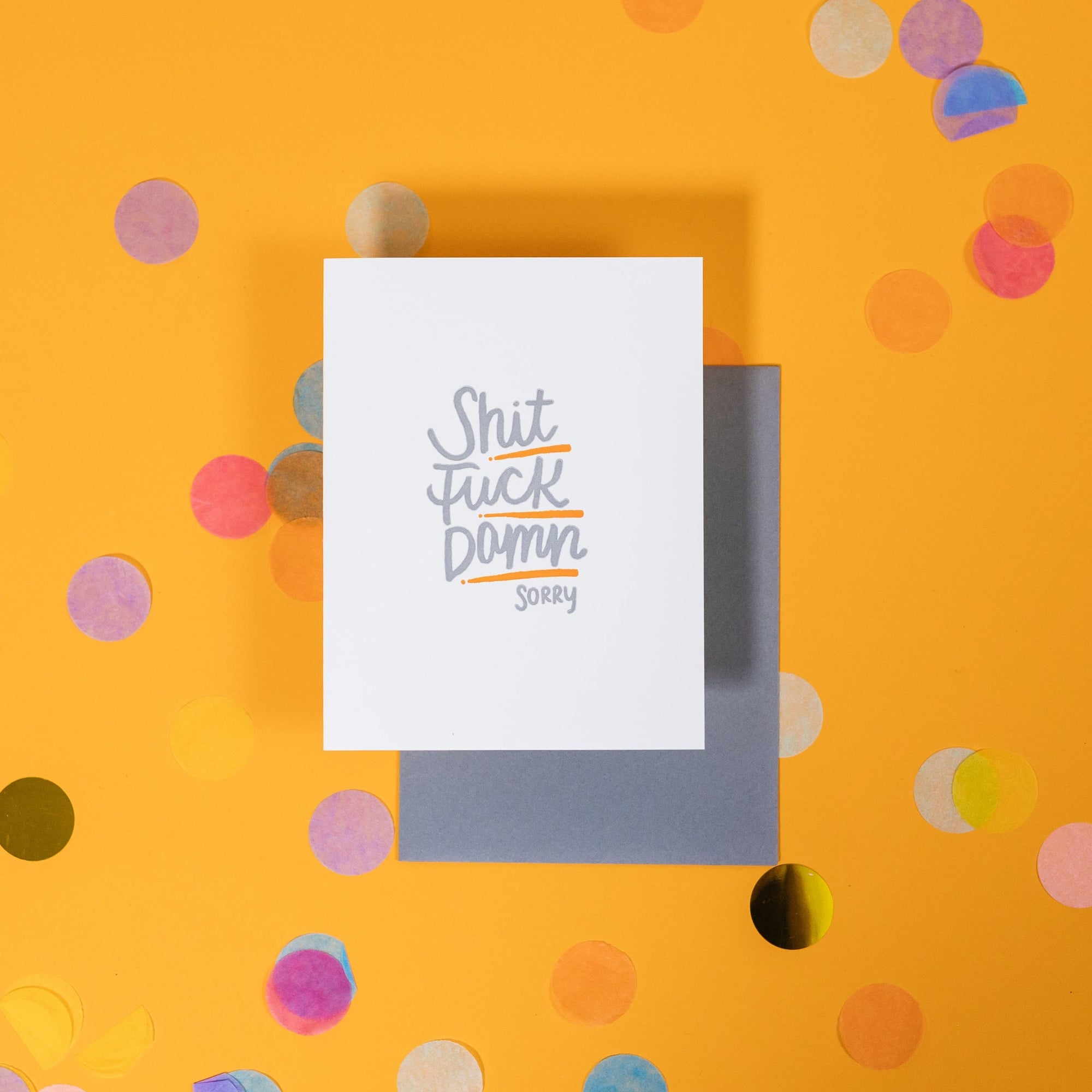 On a sunny mustard background is a greeting card and envelope with big, colorful confetti scattered around. The white greeting card says "Shit Fuck Damn Sorry" in handwritten grey lettering and underlined in orange. The dark grey envelope sits under the card. 