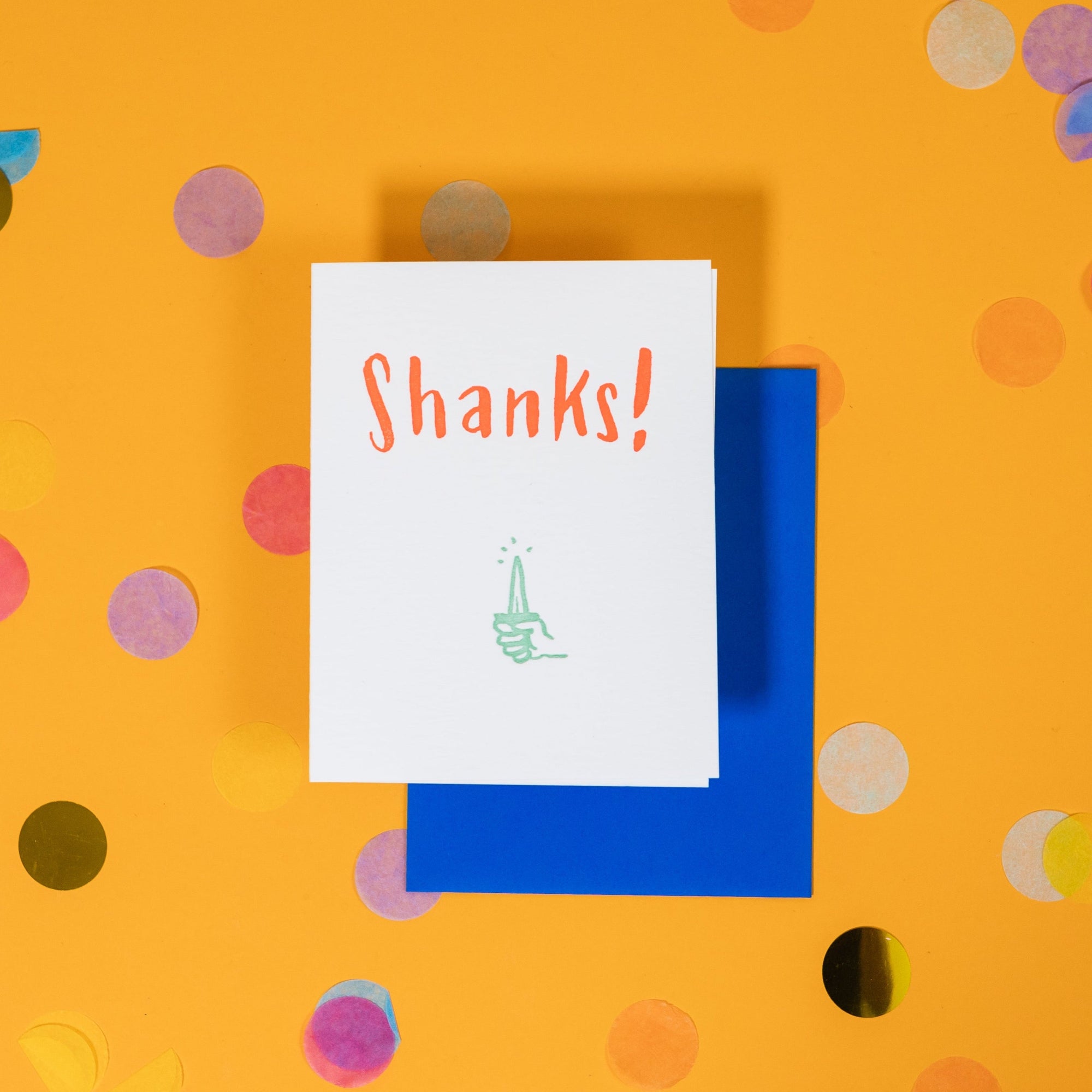On a sunny mustard background is a greeting card and envelope with big, colorful confetti scattered around. The white greeting card has an illustration of a hand holding a shank in mint and it says "Shanks!" in handwritten orange lettering. The neon blue envelope sits under the card. 