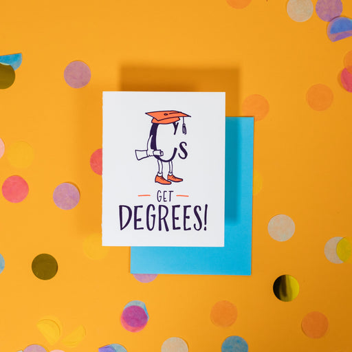 On a sunny mustard background is a greeting card and envelope with big, colorful confetti scattered around. The white greeting card has an illustration of the letter "C" with a graduation cap in orange on it and a hand holding a rolled up paper with legs and orange shoes that says "Cs Get Degrees" in handwritten black font. The turquoise blue envelope sits under the card. 