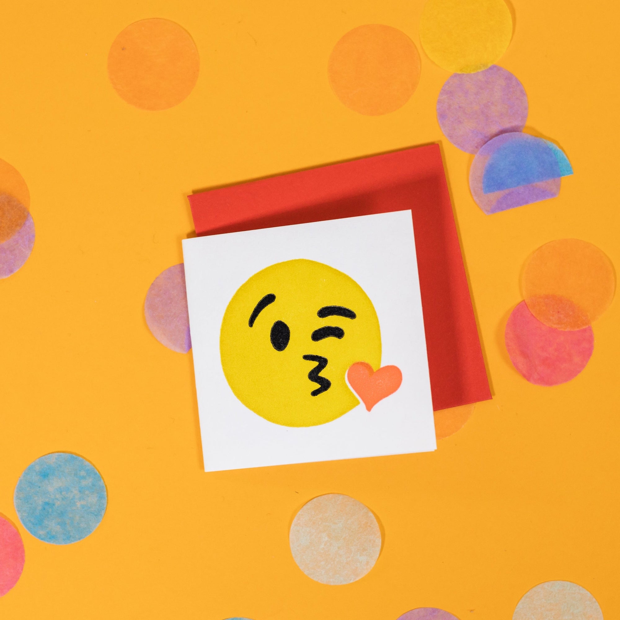 On a sunny mustard background is a mini greeting card and envelope surrounded by big, colorful confetti. The white mini card has a letterpress illustration of a kissy Emoji face winking. It is a yellow face with a red heart kiss. The envelope is a red color. 2.5"x2.5"