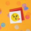 On a sunny mustard background is a mini greeting card and envelope surrounded by big, colorful confetti. The white mini card has a letterpress illustration of a kissy Emoji face winking. It is a yellow face with a red heart kiss. The envelope is a red color. 2.5"x2.5"