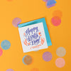 On a sunny mustard background is a mini greeting card and envelope surrounded by big, colorful confetti. The white mini card says "OMG THANK YOU!" in a coral orange handwritten serif lettering in letterpress. The envelope is a goldcolor. 2.5"x2.5"