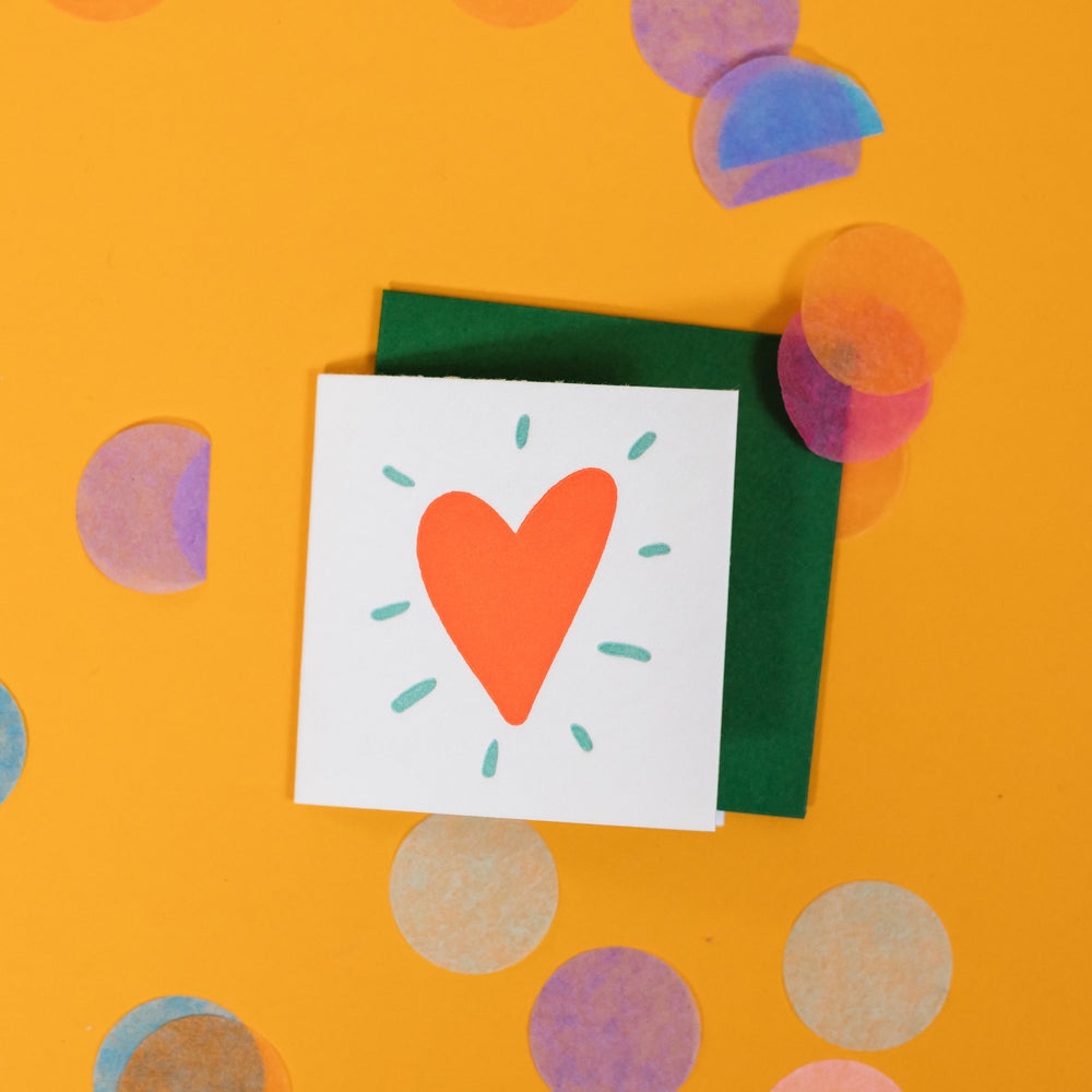 On a sunny mustard background is a mini greeting card and envelope surrounded by big, colorful confetti. The white mini card has an illustration of a red heart with mint blue bursts around it in letterpress. The envelope is a dark green color. 2.5"x2.5"