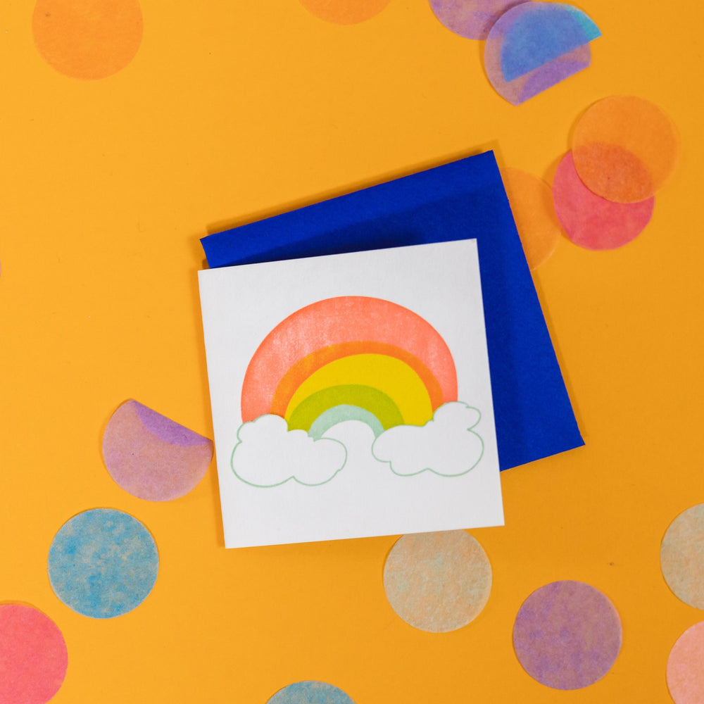 On a sunny mustard background is a mini greeting card and envelope surrounded by big, colorful confetti. The white mini card has a letterpress illustration of a rainbow and clouds. The rainbow is in orange-red, yellow, green and mint blue with white clouds outlined in mint blue. The envelope is a neon blue color. 2.5"x2.5"