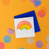 On a sunny mustard background is a mini greeting card and envelope surrounded by big, colorful confetti. The white mini card has a letterpress illustration of a rainbow and clouds. The rainbow is in orange-red, yellow, green and mint blue with white clouds outlined in mint blue. The envelope is a neon blue color. 2.5"x2.5"