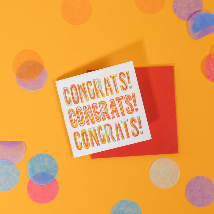 On a sunny mustard background is a mini greeting card and envelope surrounded by big, colorful confetti. The white mini card says "CONGRATS! CONGRATS! CONGRATS!" in a block, handwrittenlettering in letterpress. There are colors of orange, light blue and sunny mustard. The envelope is a red color. 2.5"x2.5"