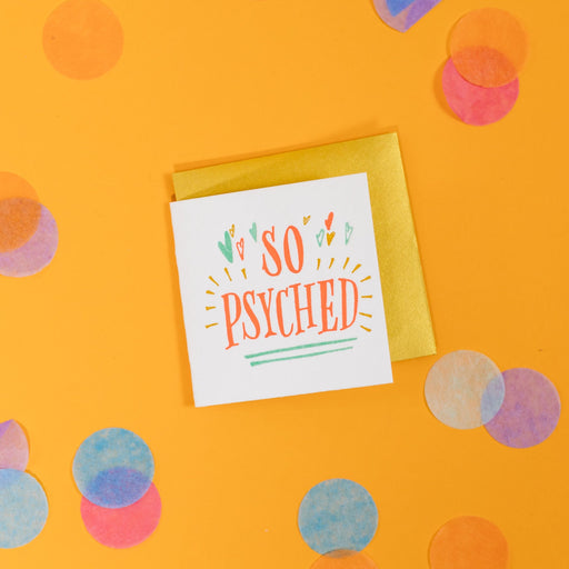 On a sunny mustard background is a mini greeting card and envelope surrounded by big, colorful confetti. The white mini card says "SO PSYCHED" in a coral orange handwritten serif lettering in letterpress. It has mustard yellow bursts around the word "PSYCHED" and mint blue, coral orange, and mint blue hearts at the top. The word "PSYCHED" is underlined twice in mint blue. The envelope is a gold color. 2.5"x2.5"