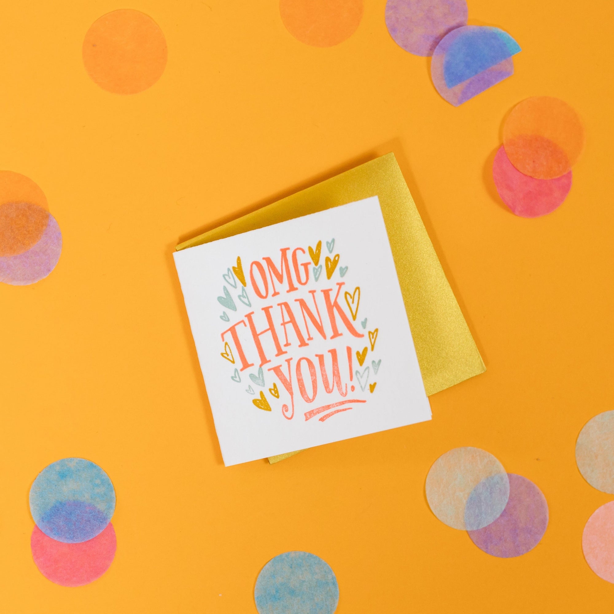 On a sunny mustard background is a mini greeting card and envelope surrounded by big, colorful confetti. The white mini card says "OMG THANK YOU!" in a coral orange handwritten serif lettering in letterpress. The envelope is a gold color. 2.5"x2.5"