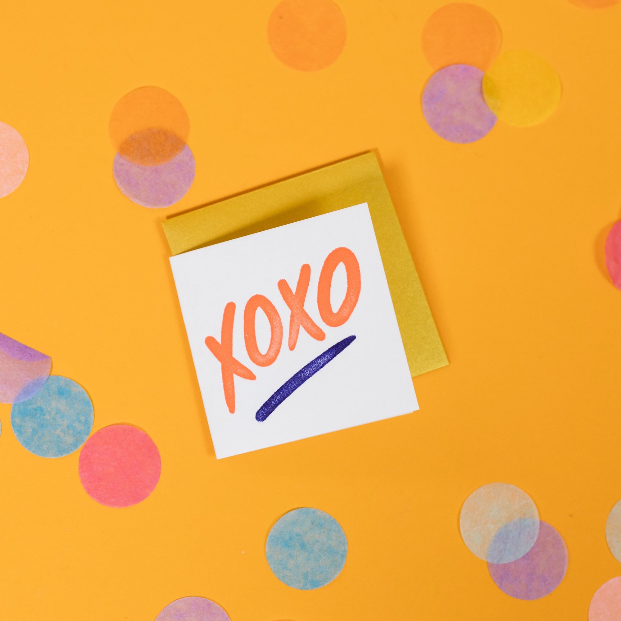 On a sunny mustard background is a mini greeting card and envelope surrounded by big, colorful confetti. The white mini card says "XOXO" in a coral orange handwritten marker lettering in letterpress with a purple underline. The envelope is a gold color. 2.5"x2.5"
