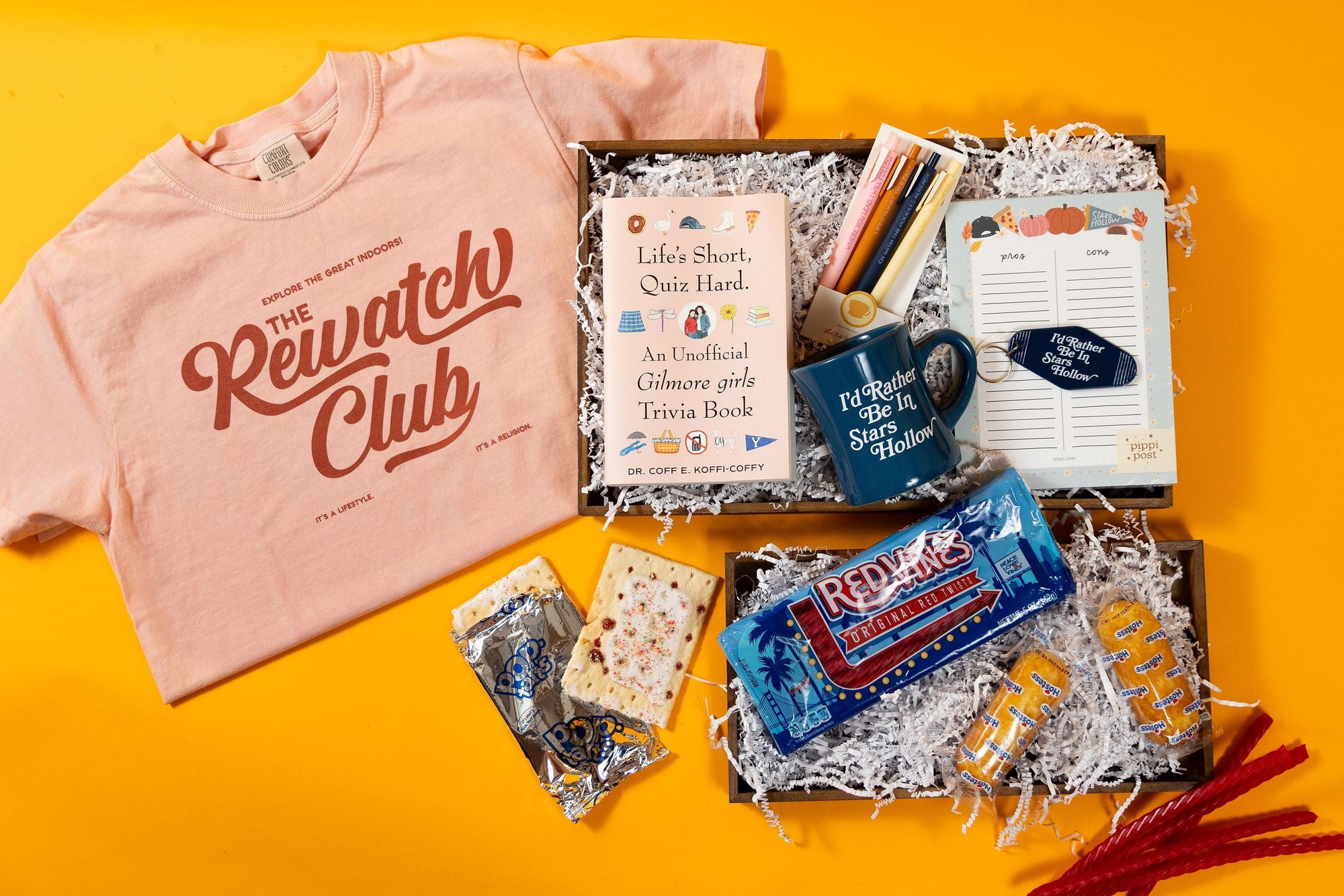 A curated gift box flat lay on a bright yellow background, featuring a light pink crewneck t-shirt with 'The Rework Club' printed on it, a book, a blue ceramic mug, a notebook, candy, crackers, and red pipe cleaners arranged in white paper shred.