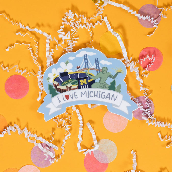 On a sunny mustard background sits a sticker with white crinkle and big, colorful confetti scattered around. This light blue sticker has an illustration of the University of Michigan stadium, pine trees, apple blossom flowers, Mackinac Bridge and the Spirit of Detroit statue. It is colorful with blues, yellows, greens and whites and there is a white banner under the illustration that says "I Love Michigan" in handwritten blue lettering. The "Love" has a red heart in place of the letter "O." 2"-3"