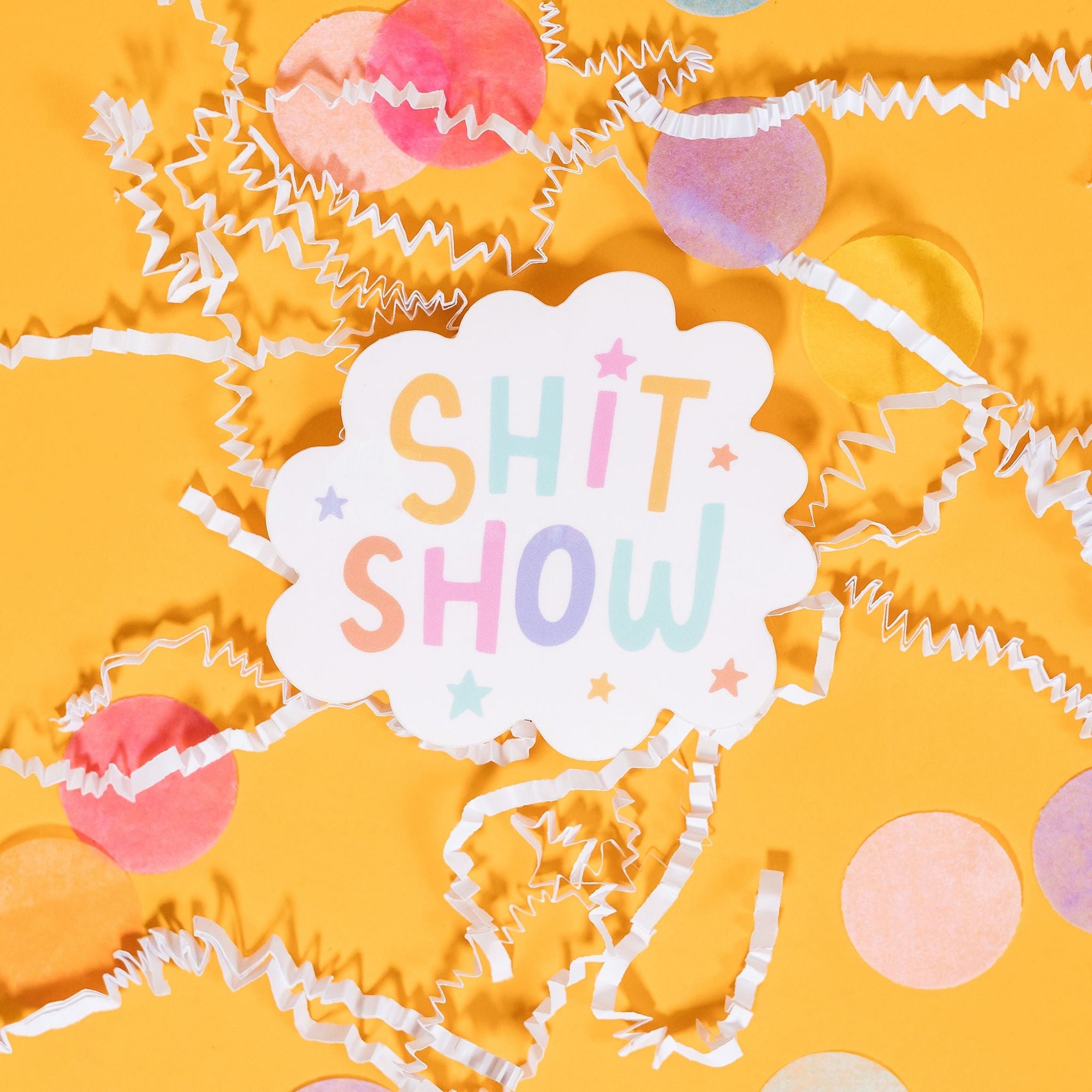 On a sunny mustard background sits a sticker with white crinkle and big, colorful confetti scattered around This white scallop shaped sticker has illustrated stars in pastel colors of purple, pink, orange, mustard yellow, aqua blue, and purple. It says "SHIT SHOW" in handwritten block lettering in pastel colors of purple, pink, orange, mustard yellow, aqua blue, and purple. 2"-3"
