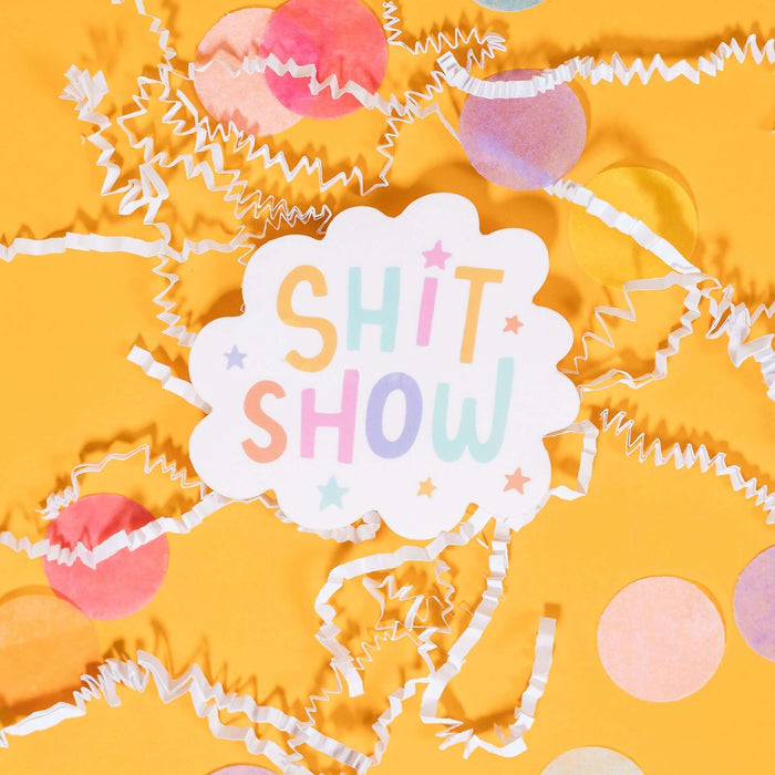On a sunny mustard background sits a sticker with white crinkle and big, colorful confetti scattered around This white scallop shaped sticker has illustrated stars in pastel colors of purple, pink, orange, mustard yellow, aqua blue, and purple. It says "SHIT SHOW" in handwritten block lettering in pastel colors of purple, pink, orange, mustard yellow, aqua blue, and purple. 2"-3"