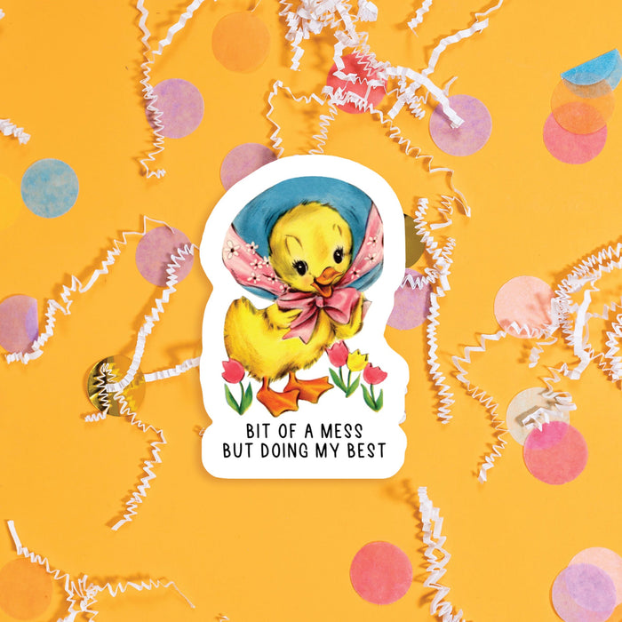 On a sunny mustard background sits a sticker with colorful confetti and white crinkle scattered around. This vintage sticker has an illustration of a darling yellow duckling with a wide-brim blue hat and pink ribbon tied around the neck. It says "BIT OF A MESS BUT DOING MY BEST" in black, handwritten all caps lettering. Approximately 3".