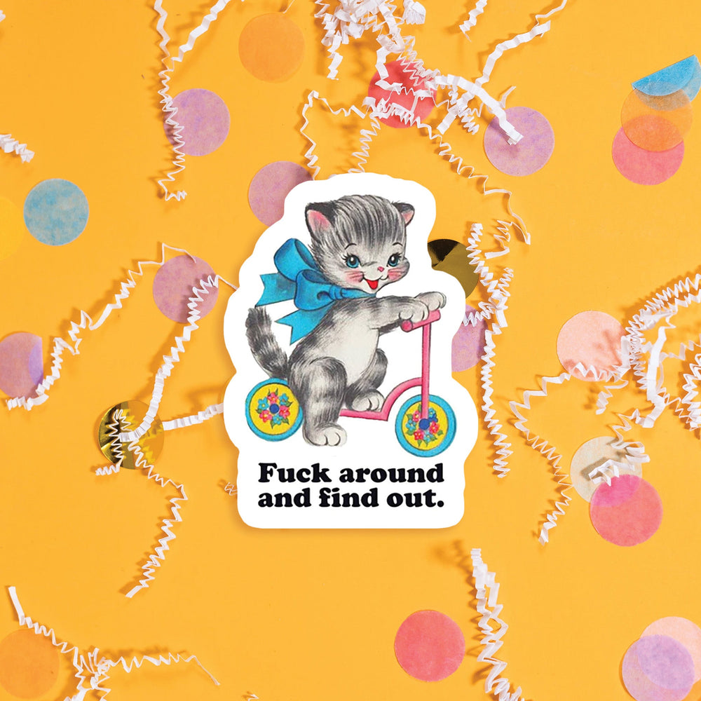 On a sunny mustard background sits a sticker with colorful confetti and white crinkle scattered around. This vintage sticker has an illustration of a darling white, gray, and black kitten on a pink, blue, and yellow scooter with a blue ribbon tied around it's neck. It says "Fuck around and find out" in black, thick serif font. Approximately 3".