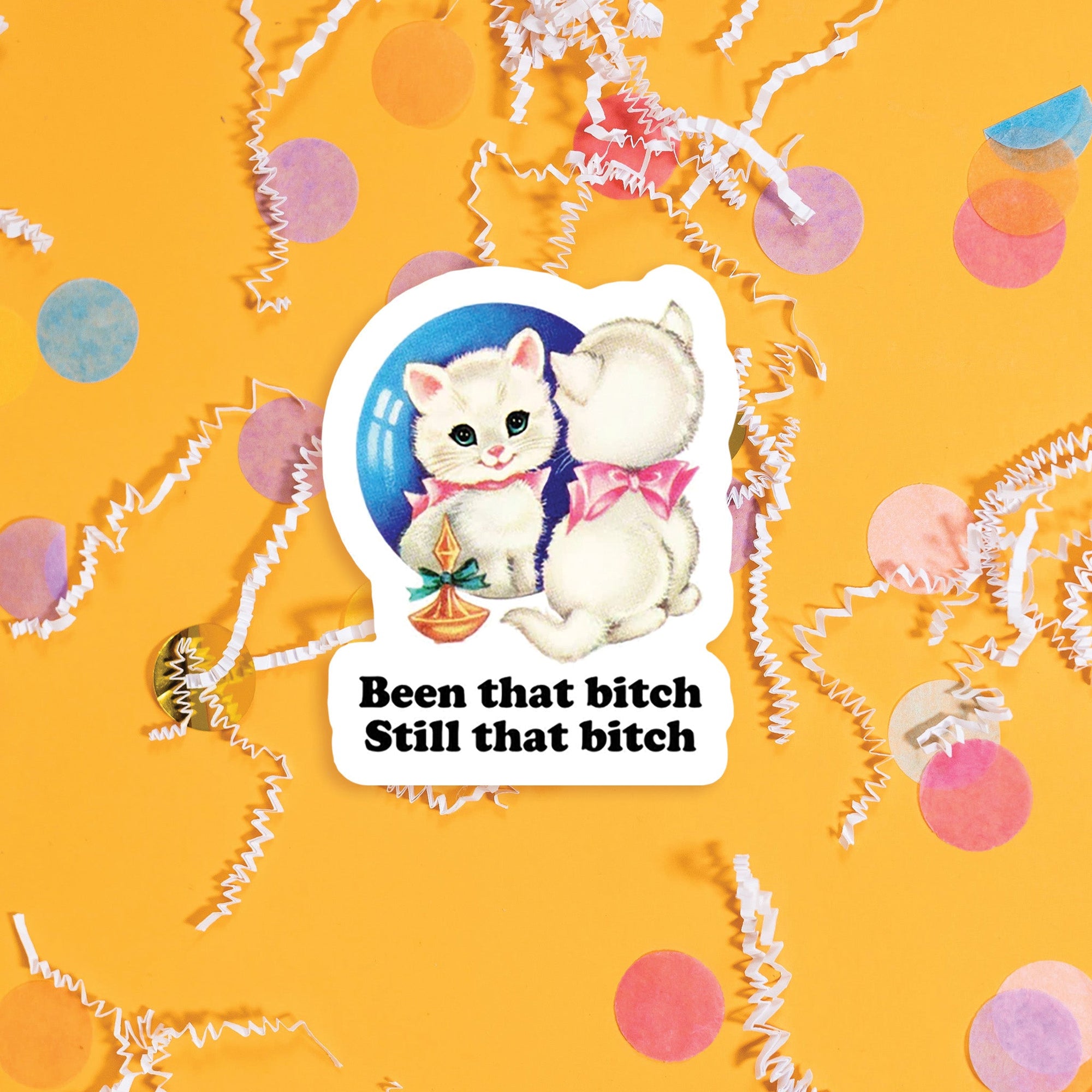 On a sunny mustard background sits a sticker with colorful confetti and white crinkle scattered around. This vintage sticker has an illustration of a darling white kitten staring at itself in a mirror. It says "Been that bitch Still that bitch" in black, thick serif font. Approximately 3".