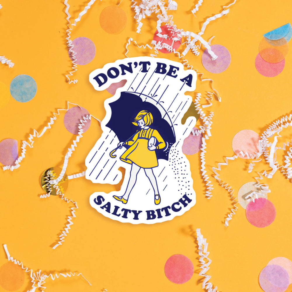On a sunny mustard background sits a sticker with colorful confetti and white crinkle scattered around. This vintage sticker has an illustration of a young girl under an umbrella holding a salt shaker that is dumping salt. It is raining. The colors are navy and sunny mustard. It says "DON'T BE A SALTY BITCH" in navy, all caps thick serif font. Approximately 3".