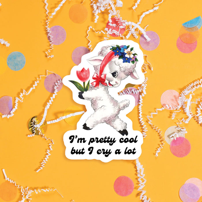 On a sunny mustard background sits a sticker with colorful confetti and white crinkle scattered around. This vintage sticker has an illustration of a cute white lamb holding a red tulip and a garland of flowers on it's head tied with a red ribbon around it's neck. It says "I'm pretty cool but I cry a lot" in black, handdwritten lettering. Approximately 3".