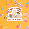 On a sunny mustard background sits a sticker with colorful confetti and white crinkle scattered around. This vintage sticker has an illustration of an open book with a sun, rainbow, cloud, and flowers. It says "TAKE A LOOK IT'S IN A BOOK" in brown, all caps groovy font. Approximately 3".