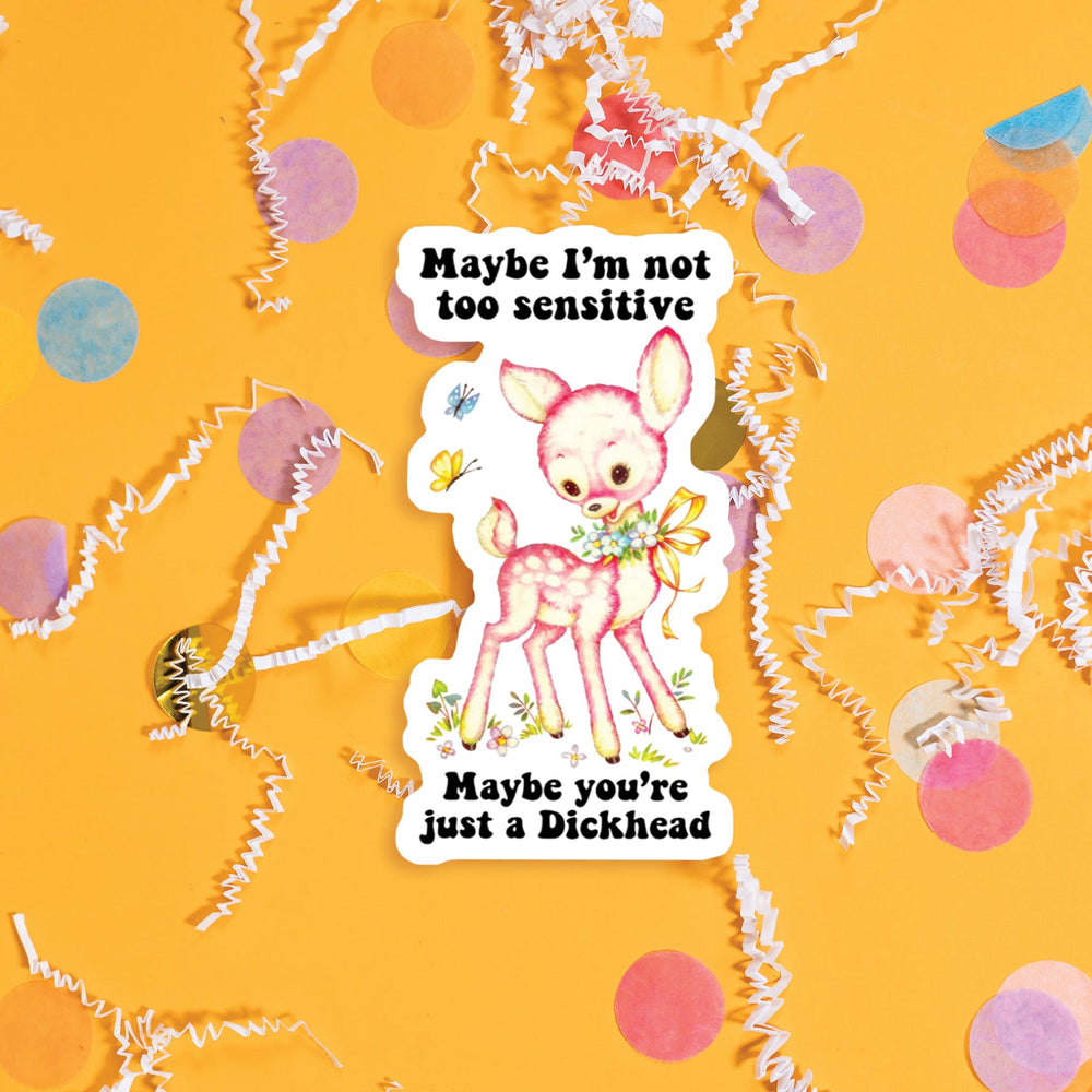 On a sunny mustard background sits a sticker with colorful confetti and white crinkle scattered around. This vintage sticker has an illustration of a cute pink deer with a floral garland tied with a yellow ribbon around the neck. It says "Maybe I'm not too sensitive Maybe you're just a Dickhead" in black, thick serif font. Approximately 3".