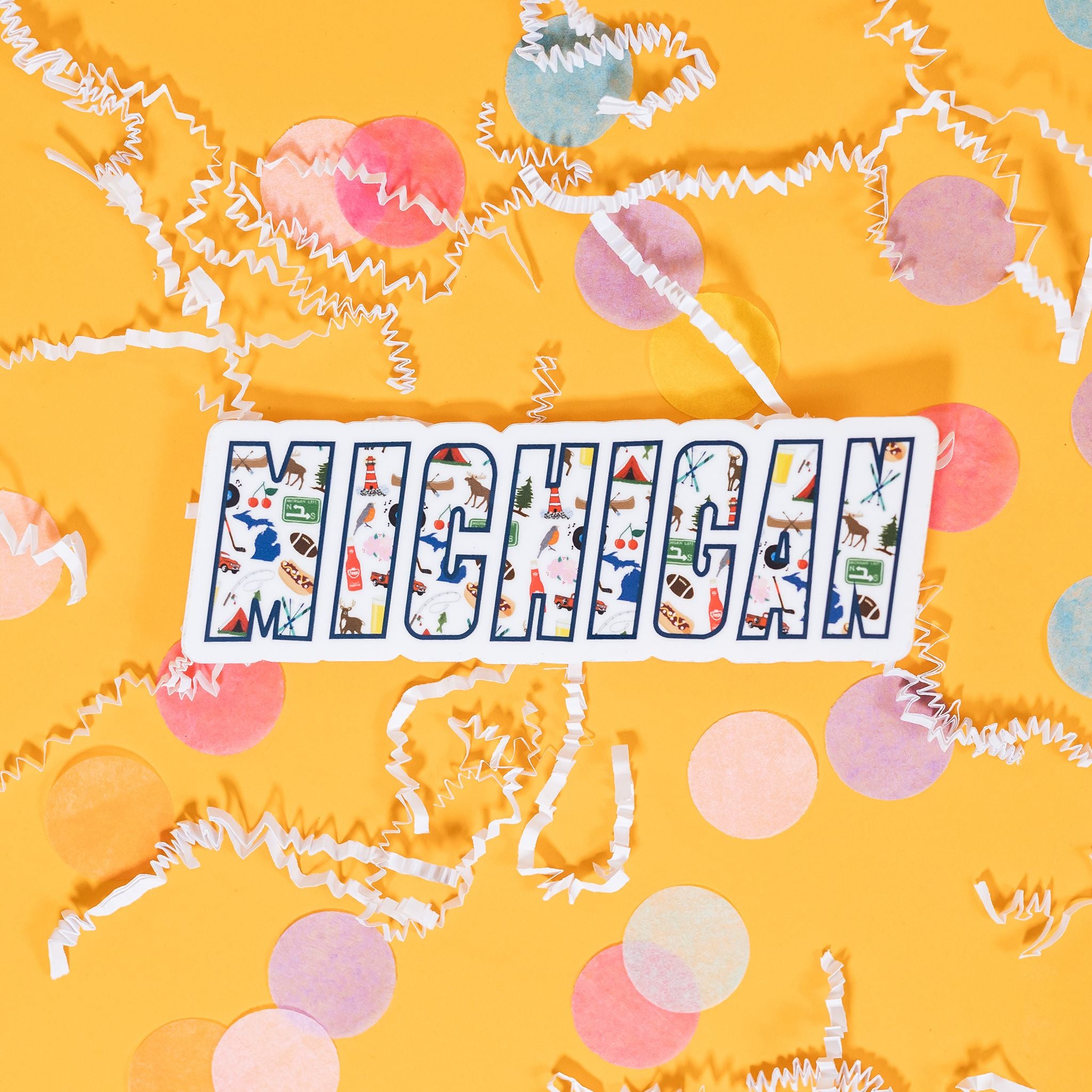 On a sunny mustard background sits a sticker with white crinkle and big, colorful confetti scattered around. This white sticker says "MICHIGAN" in a navy outline will illustrations of icons inside the word. The colorful illustrations are of Michigan favorites (Motown, Vernors, Faygo, coney dogs, the Michigan left) as well as activities, symbols, and landmarks. 3"-4"