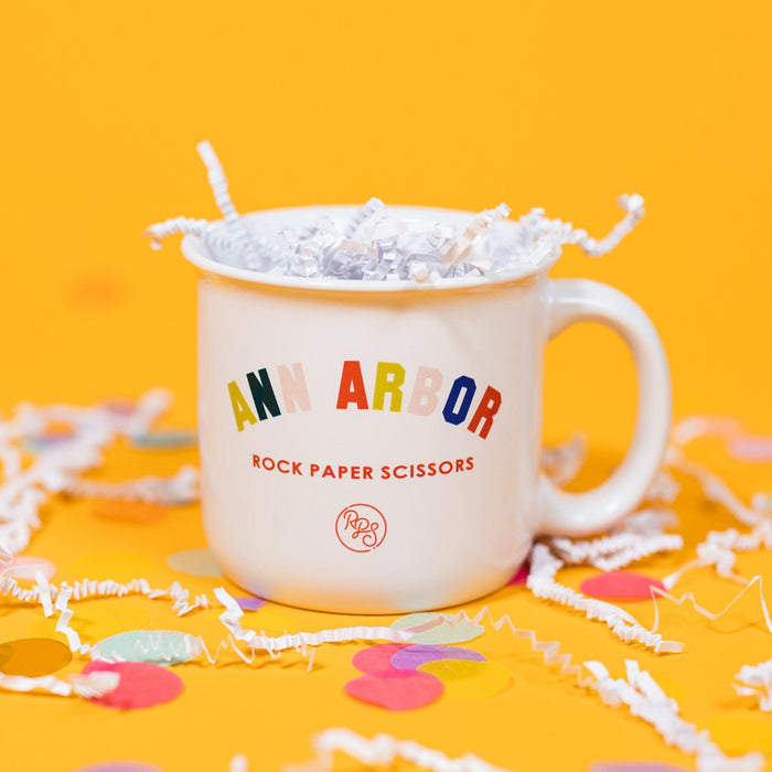 On a sunny mustard background sits a mug with white crinkle and big, colorful confetti scattered around. The white mug has a color collegiate font that says "ANN ARBOR" and under it says "ROCK PAPER SCISSORS" in a red, all caps block font. Under that is a circular, red "RPS" logo in handwritten script lettering.