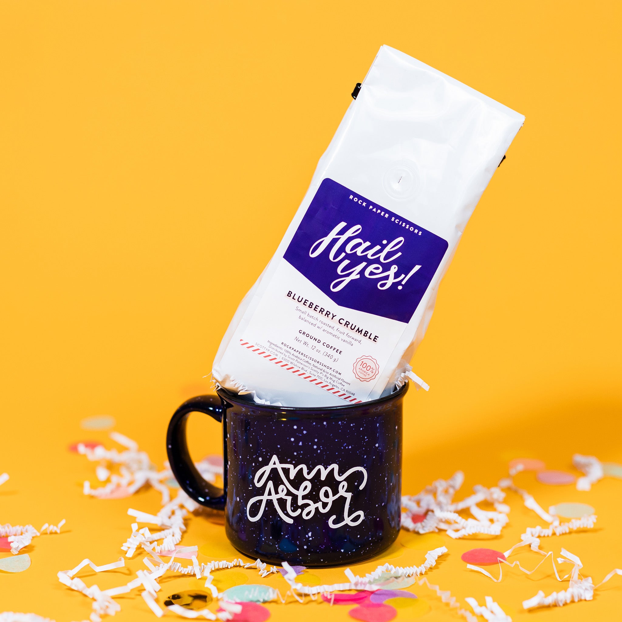 On a sunny mustard background sits a mug and a bag of coffee with white crinkle and big, colorful confetti scattered around.  The navy campfire mug has white specks and says "Ann Arbor" in a white, handwritten script lettering font. The white bag of coffee sits atop the mug. The coffee is Rock Paper Scissors 'Hail Yes!' in white hand lettering on a brilliant blue label. It is 'BLUEBERRY CRUMBLE GROUND COFFEE'. Small batch roasted, fruit forward, balanced w/ aromatic vanilla. Net Wt. 12 oz. (340g)