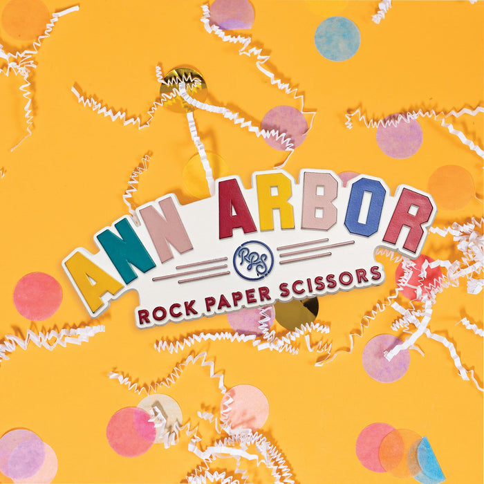 On a sunny mustard background sits a magnet with white crinkle and big, colorful confetti scattered around. This colorful 3-D magnet says "ANN ARBOR" in colors of mustard yellow, teal, pink, red, and blue. It has the RPS logo and under it says "ROCK PAPER SCISSORS" in red. 