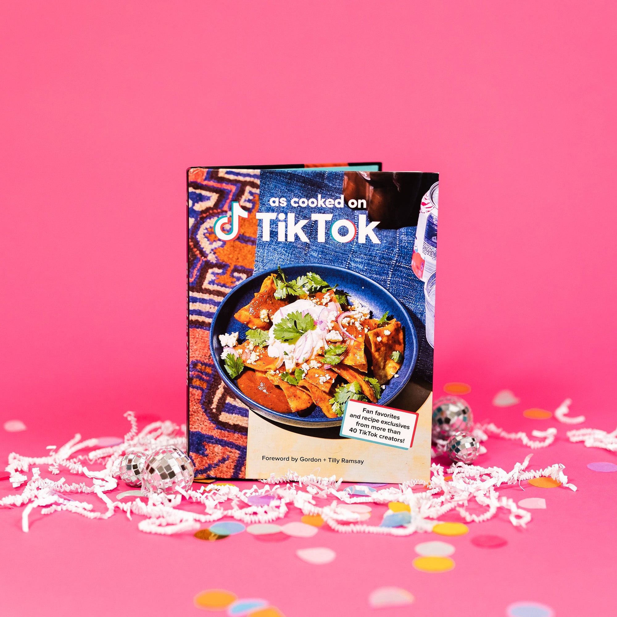 On hot pink background sits a book with white crinkle and big, colorful confetti. There are mini disco balls. This book has a picture of a blue bowl filled with tortilla chips, cilantro, white cheese and onions. It says "as cooked on TikTok" in a white, block font. In a white box it says "Fan favorites and recipe exclusives from more than 40 TikTok creators!" with a red and turquoise border. It also says "Foreword by Gordon + Tilly Ramsay" in black, block font.