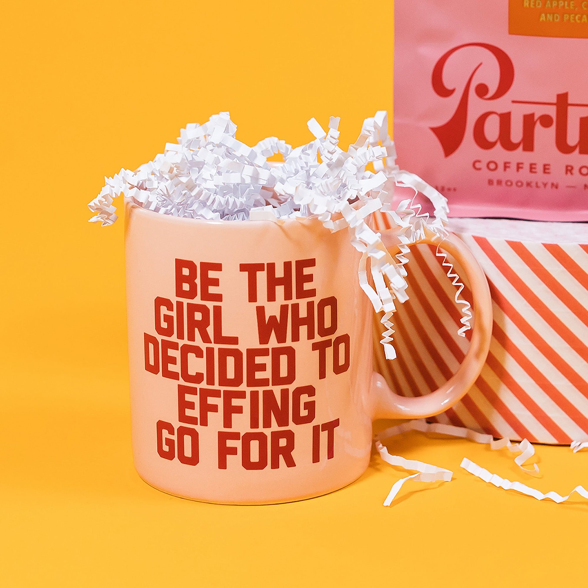 On a sunny mustard background sits a mug with white crinkle in it. The light pink mug says "BE THE GIRL WHO DECIDED TO EFFING GO FOR IT" in red, all caps block font.