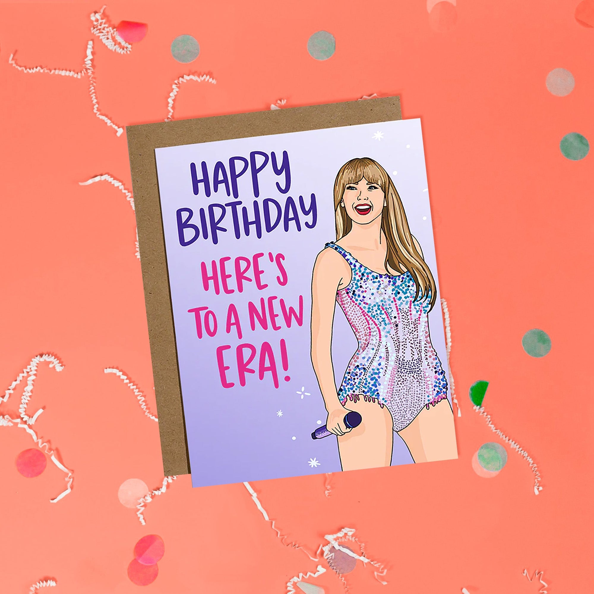 On a coral pink background sits a card with white crinkle and big, colorful confetti scattered around. This Taylor Swift inspired card has different shades of lavender. There is an illustration of Taylor Swift and it says "HAPPY BIRTHDAY HERE'S TO A NEW ERA!" in purple and pink lettering. It has a kraft envelope.