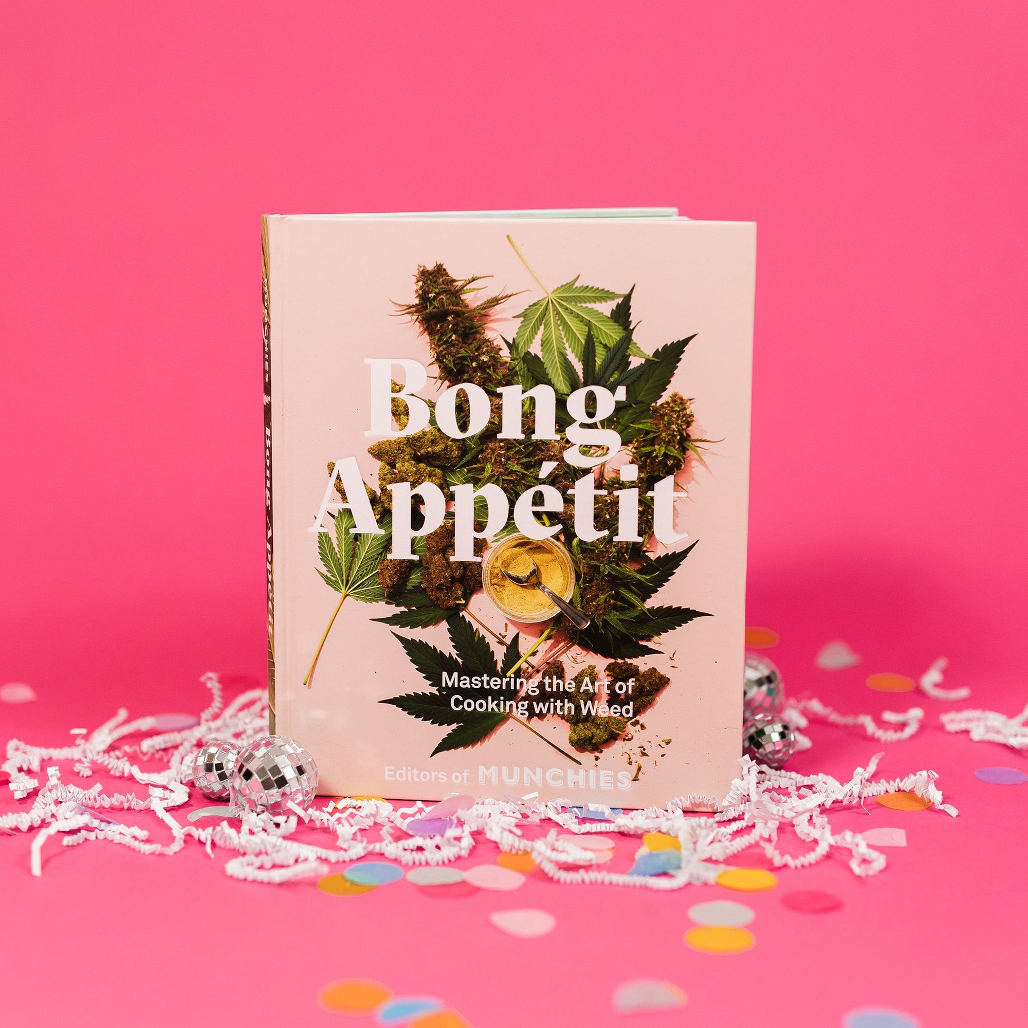 hot pink background sits a light pink book with white crinkle and big, colorful confetti. There are mini disco balls. It has a picture of marijuana leaves, marijuana buds, a jar of powdered marijana with a spoon and it says "Bong Appetit" in white, bold serif lettering. It also says at the bottom "Mastering the Art of Cooking with Weed" in white, block lettering. It is by the Editors of MUNCHIES.