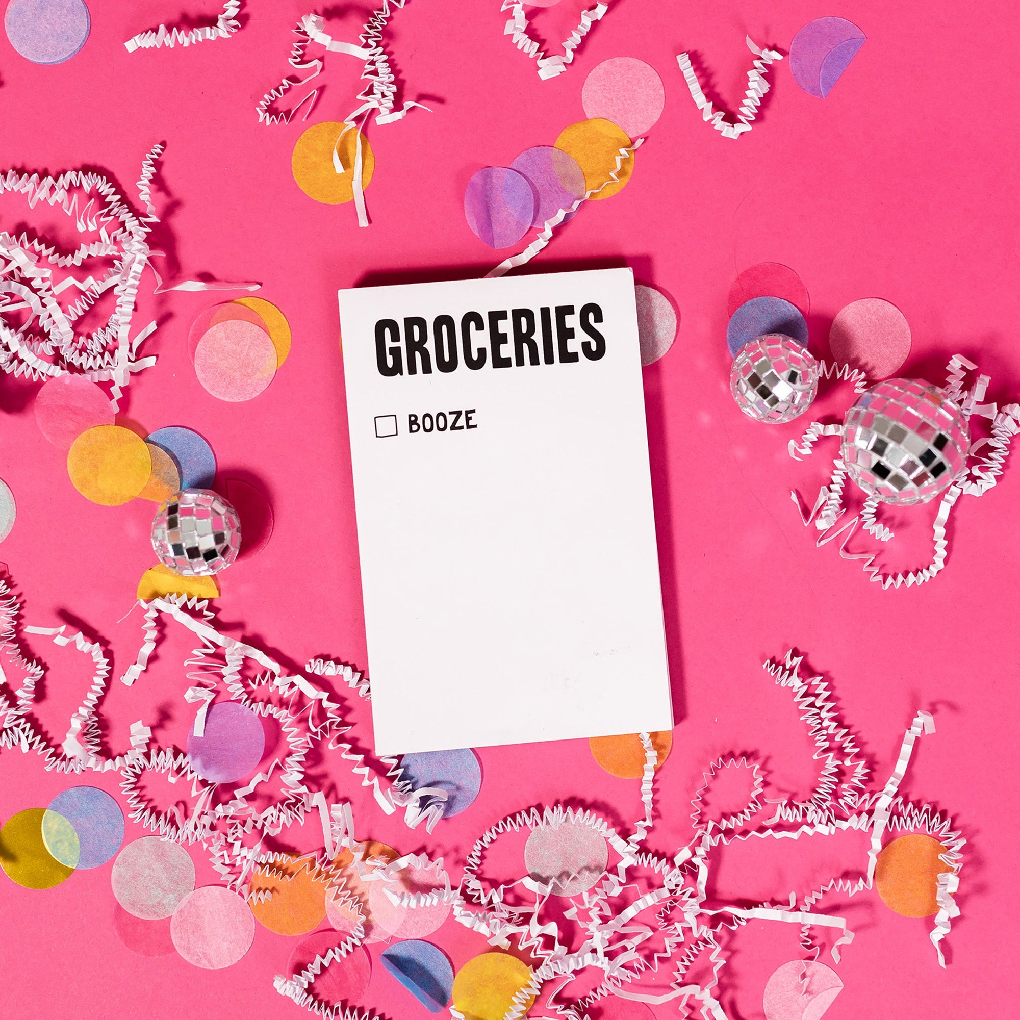 On a hot pink background sits a notepad with white crinkle and big, colorful confetti scattered around. There are mini disco balls. There is a white notepad with "GROCERIES" in black, all caps lettering at the top. Under it is a check box with the word "BOOZE" next to it in black, all caps lettering.