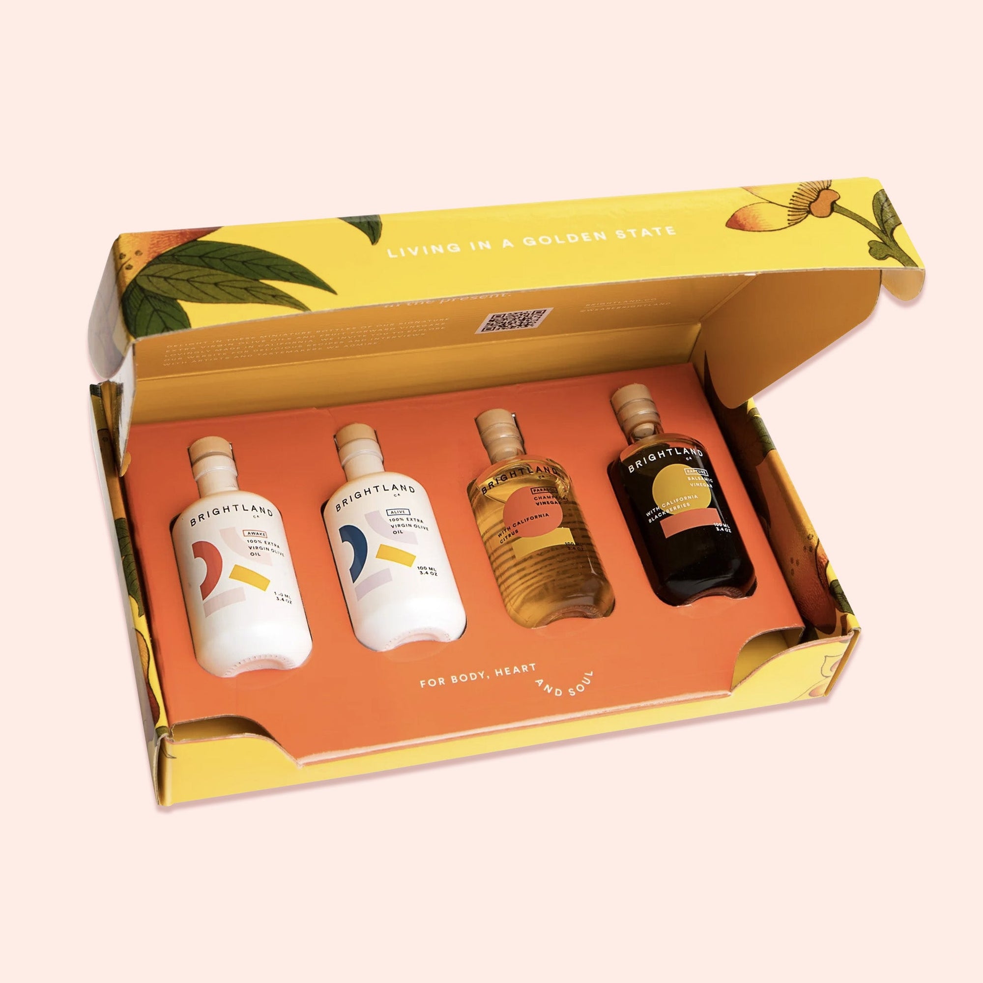 On a light pink background sits an open box of bottles. This yellow box has colorful illustrations of flowers and leaves and the inside is orange with two white bottles and two clear bottles. They are bottles of olive oils and vinegars. It has white lettering that says "LIVING IN A GOLDEN STATE" and "FOR BODY, HEART AND SOUL."