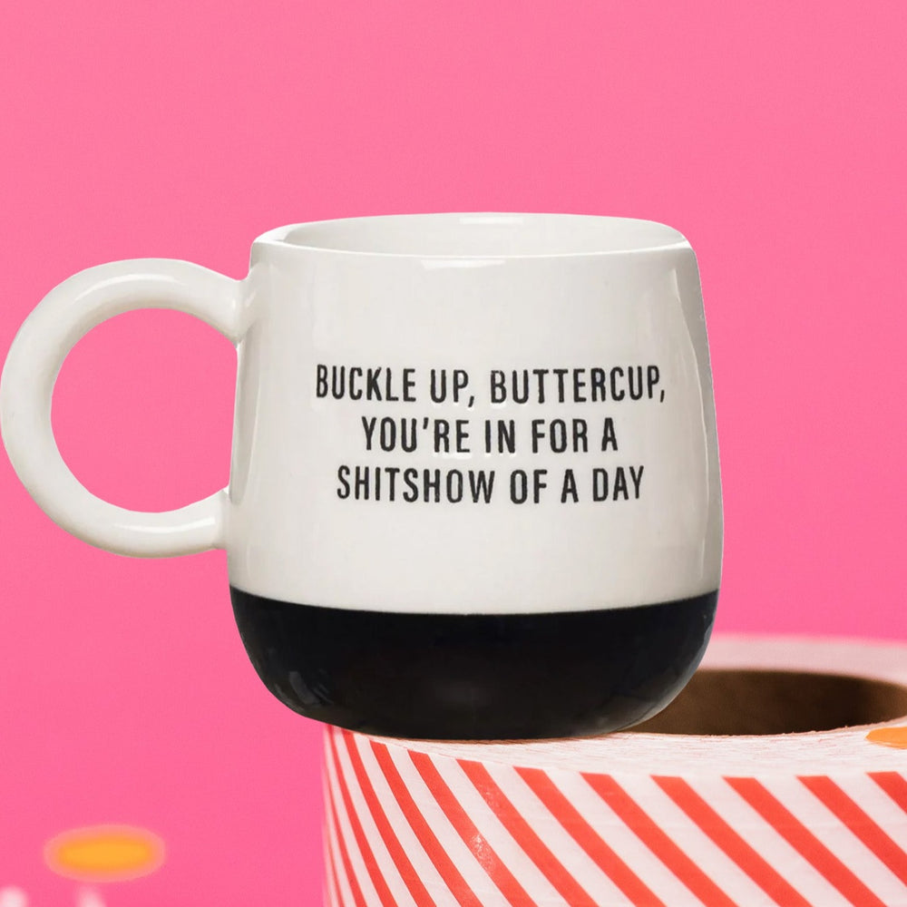 On a hot pink background sits a mug on a red and white striped packing tape with white crinkle and big, colorful confetti. The mug is half white and half black. On the white part it says "BUCKLE UP, BUTTERCUP, YOU'RE IN FOR A SHITSHOW OF A DAY" in a black, all caps block font.
