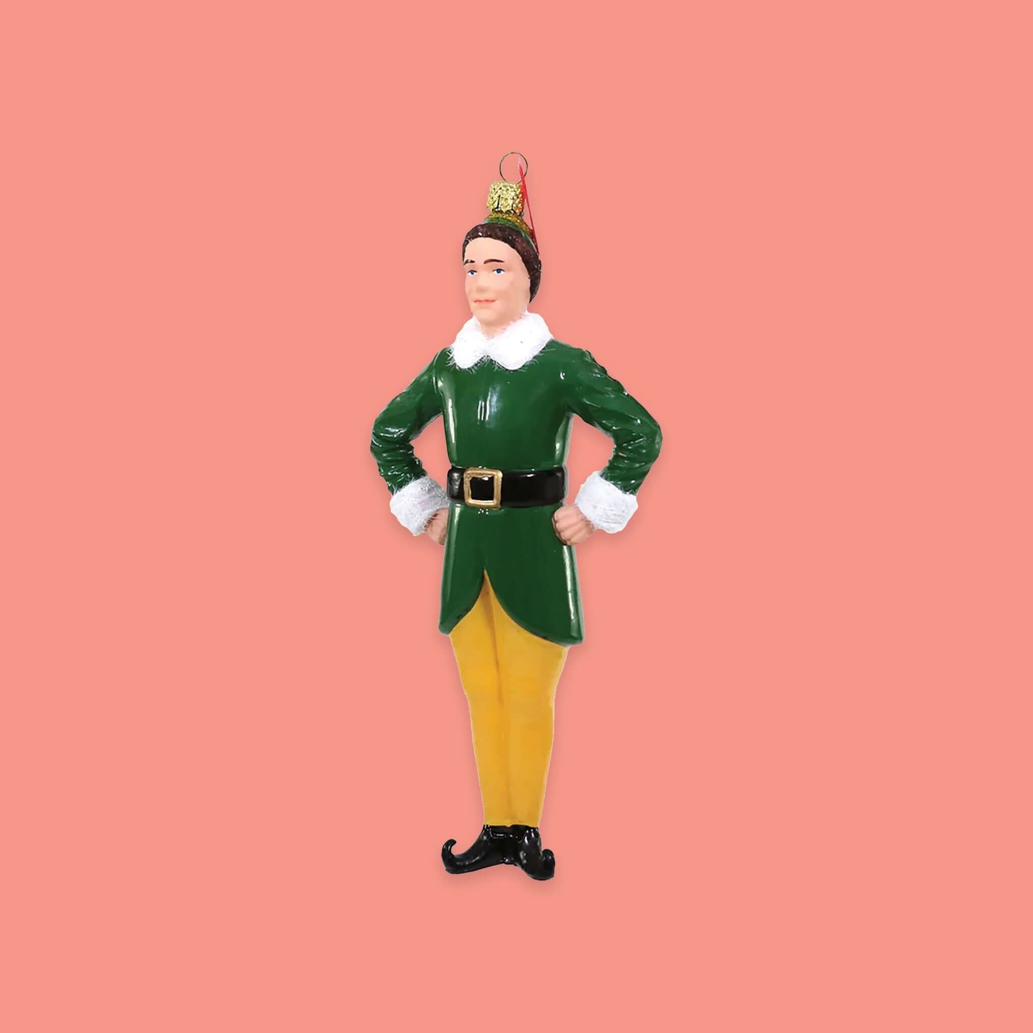 On a coral pink background sits an ornament. This Buddy the Elf inspired ceramic ornament is of Buddy wearing the famous green coat with white fur collar and cuffs and yellow pants with black elf shoes. 