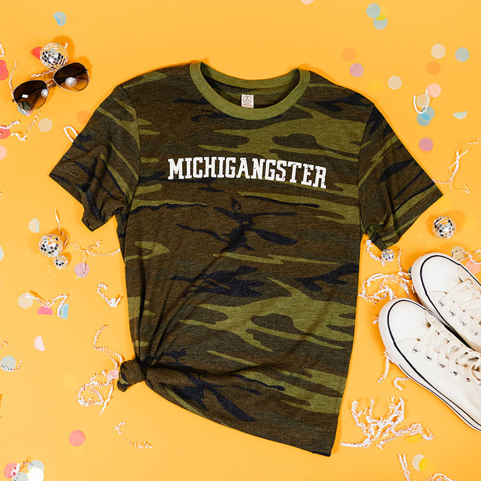On a sunny mustard background sits a t-shirt with white crinkle and big, colorful confetti scattered around. There are mini disco balls, sunglasses, and a pair of white sneakers. This camo tee features "MICHIGANGSTER" in a distressed, varsity-style white print.