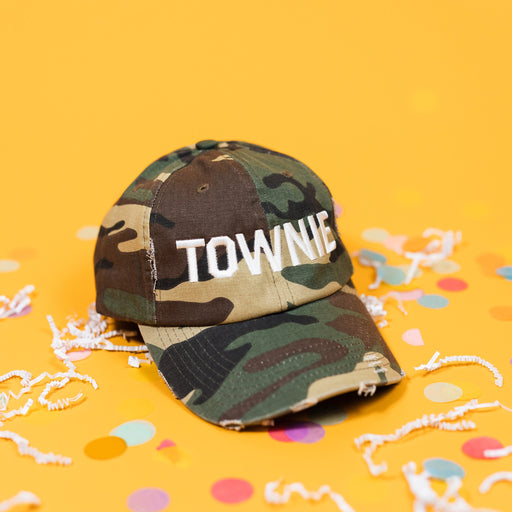 On a sunny mustard background sits a camoflauge hat with white crinkle and big, colorful confetti scattered around. The hat is distressed with bold, white embroidered lettering that says "TOWNIE."