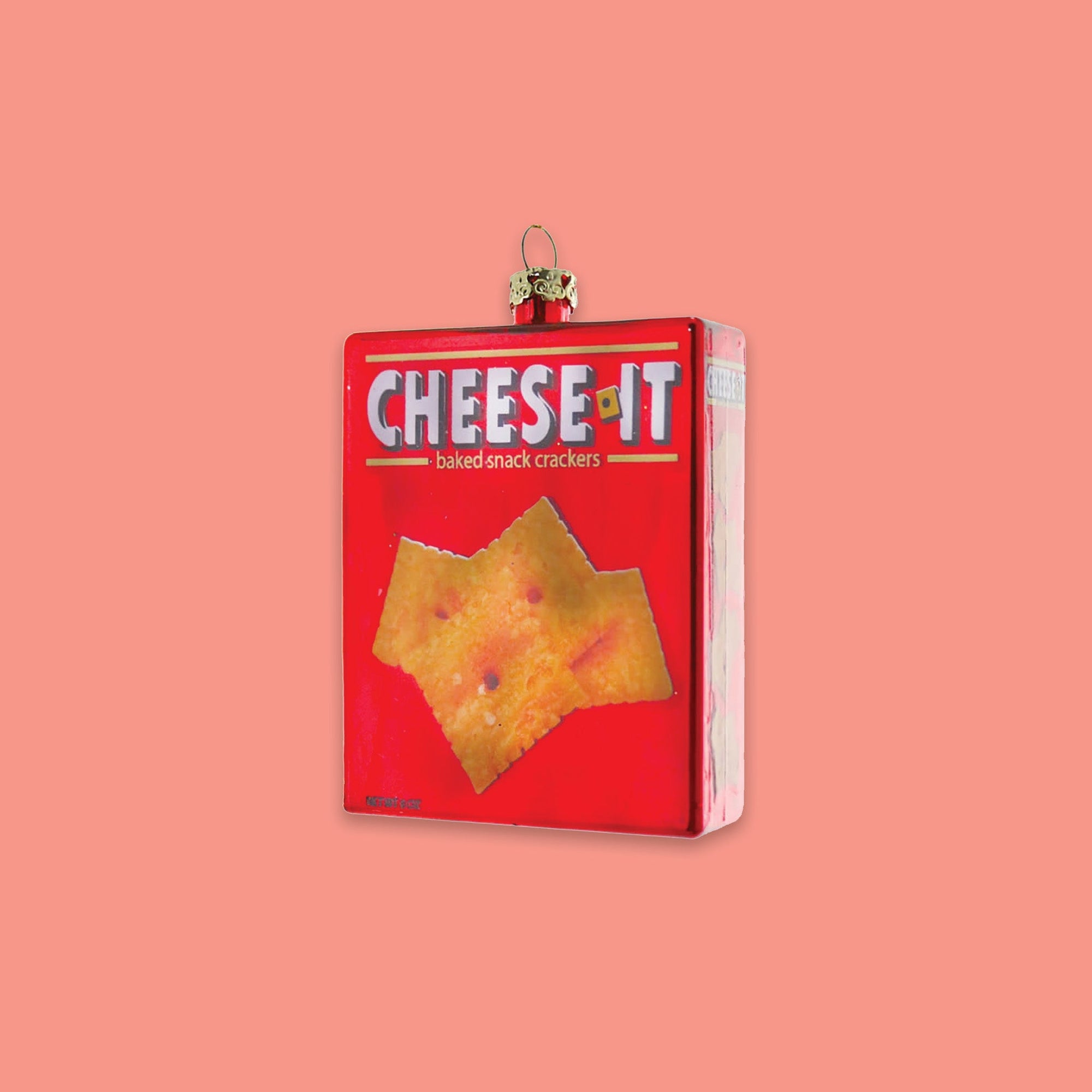 On a geometric light and medium pink background sits a box ornament. This 'CHEESE-IT' glass box ornament is red with cheese-it crackers on it. It says "baked snack crackers."