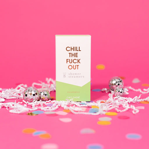 On a hot pink background sits a box with white crinkle and big, colorful confetti scattered around. There are mini disco balls. This picture is a close-up of a white and pistachio green package that says "CHILL THE FUCK OUT" in gold foil, all caps block lettering. Under it says "set of 8" and " shower steamers" in grey, lowercase serif font. At the bottom it says "eucalyptus" in gold foil, lower case serif font.