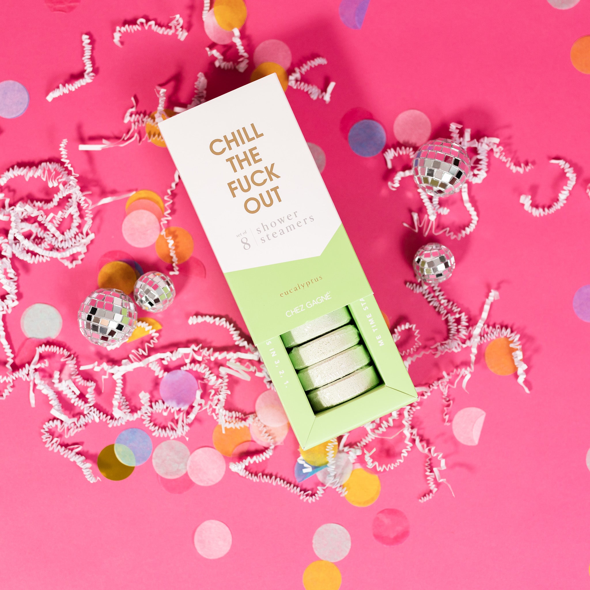 On a hot pink background sits an opened box with white crinkle and big, colorful confetti scattered around. There are mini disco balls. This picture is a close-up of a white and pistachio green package that says "CHILL THE FUCK OUT" in gold foil, all caps block lettering. Under it says "set of 8" and " shower steamers" in grey, lowercase serif font. At the bottom it says "eucalyptus" in gold foil, lower case serif font. The box is opened to reveal the pistachio green shower steamers in it. 