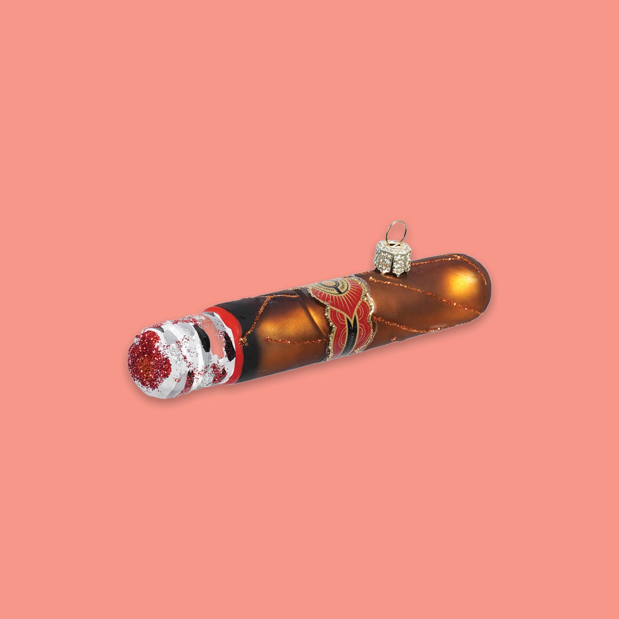 On a coral pink background sits a cigar ornament. This glass ornament looks like a real cigar with brown paper and red glitter at the end.