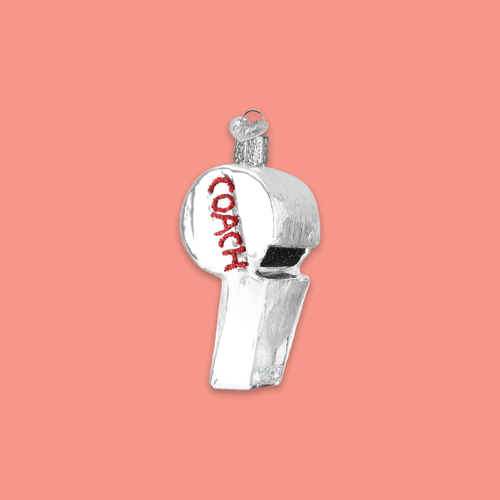 On a coral pinkbackground sits a whistle ornament. This silver glass ornament looks like a real whistle and in red glitter it says "COACH." 
