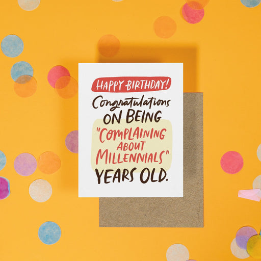 On a sunny mustard background is a greeting card and envelope with big, colorful confetti scattered around. The white greeting card says "Happy Birthday! Congratulations on being "complaining about millenials" years old." in a handwritten lettering. The colors are red, black, and light yellow. The kraft envelope sits under the card. 