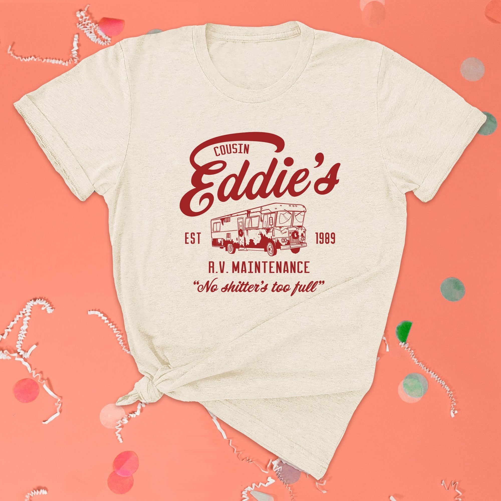 On a pink coral background sits the front of a t-shirt with white crinkle and big, colorful confetti scattered around. This Christmas Vacation Inspired crewneck ivory t-shirt has red lettering with an illustration of an old motorhome. It says "COUSIN Eddie's R.V. MAINTENANCE EST 1989" and "No shitter's too full."