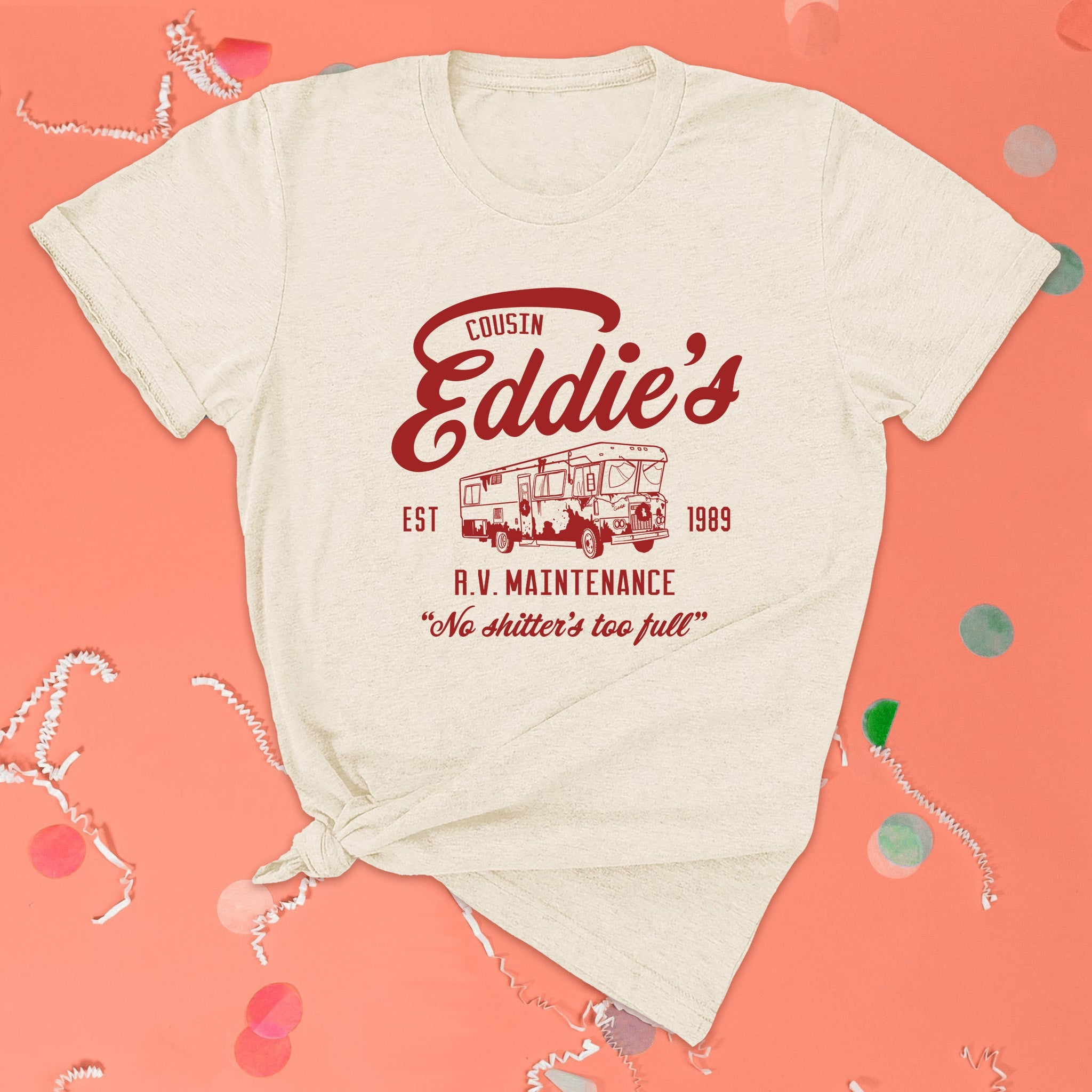 On a pink coral background sits the front of a t-shirt with white crinkle and big, colorful confetti scattered around. This Christmas Vacation Inspired crewneck ivory t-shirt has red lettering with an illustration of an old motorhome. It says "COUSIN Eddie's R.V. MAINTENANCE EST 1989" and "No shitter's too full."